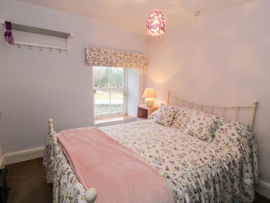 A bedroom with a floral bedspread and pillows a bedside table with a lamp and a window with a floral roman blind at Miners Cottage in Blaenau Ffestiniog
