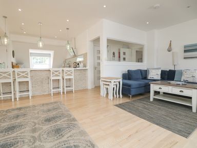 An open living room and kitchen area with a blue sofa, white stools at a breakfast bar, and wooden floor at Dawn Catcher in Falmouth
