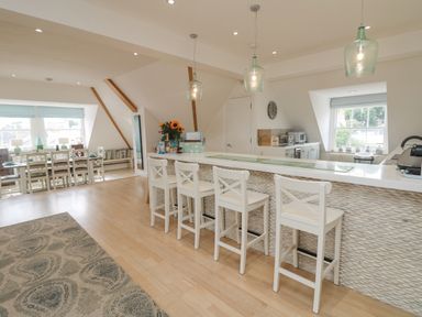 A kitchen with white chairs at a breakfast bar and a dining table with chairs near windows at Dawn Catcher in Falmouth