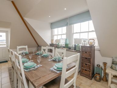 A dining room with a wooden table set with dishes and glasses near windows and storage furniture at Dawn Catcher in Falmouth