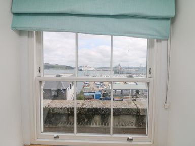 View through a window with a light blue roman blind showing a marina with boats and ships in the background at Dawn Catcher in Falmouth
