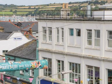 A street view with buildings including a white building and a green building with a banner and a sign that reads The Stable with boats and hills in the background at Dawn Catcher in Falmouth