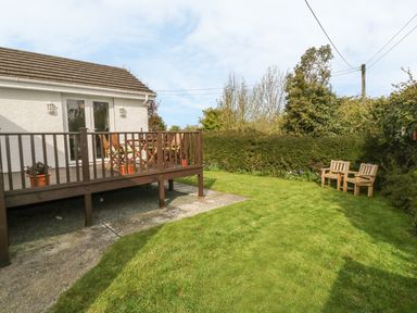 A backyard with a wooden deck and chairs next to a white house with a grassy lawn and wooden benches at Tyn Y Giat in Brynsiencyn near Beaumaris