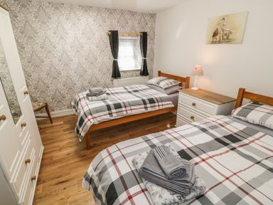 A bedroom with two single beds a bedside table with a lamp a wardrobe a chair and a window with curtains at Tyn Y Giat in Brynsiencyn near Beaumaris