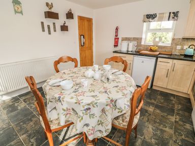 A kitchen with a round table set for tea and wooden chairs near cabinets and a window at Tyn Y Giat in Brynsiencyn near Beaumaris