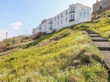 A row of white buildings on a hillside with concrete steps and green grass with small white flowers at Sennen Heights in Sennen Cove near Penzance