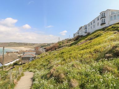 A coastal pathway with grass and white flowers beside houses on a hillside overlooking a beach at Sennen Heights in Sennen Cove near Penzance