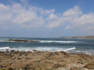 Rocky coastline with waves and distant cliffs under a partly cloudy sky at Sennen Heights in Sennen Cove near Penzance