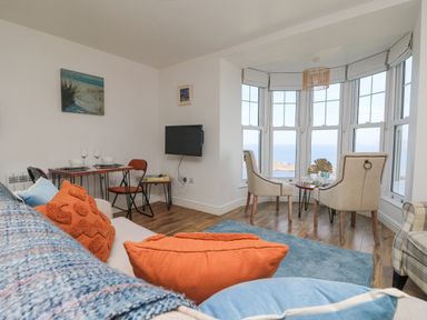 A living room with a sofa orange and blue cushions a small dining table with plates wine glasses two beige chairs by bay windows overlooking the sea at Sennen Heights in Sennen Cove near Penzance