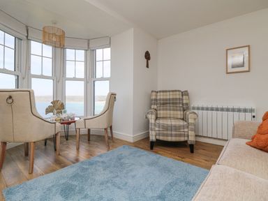 A sitting area with two beige chairs near windows overlooking water a plaid armchair and a blue rug on wooden floor at Sennen Heights in Sennen Cove near Penzance