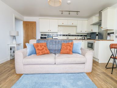 A beige sofa with orange and blue cushions in front of a white kitchen with wooden countertops and appliances at Sennen Heights in Sennen Cove near Penzance