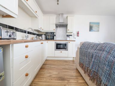 A kitchen with white cabinets wooden knobs a toaster oven microwave and stove alongside a sofa covered with a blanket at Sennen Heights in Sennen Cove near Penzance