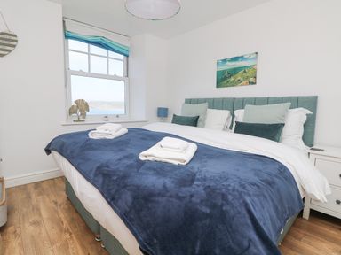 A bedroom with a double bed, blue and white bedding, bedside tables, a window with a view, and a wall picture at Sennen Heights in Sennen Cove near Penzance