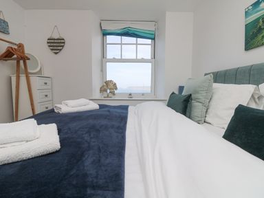 A bedroom with a bed covered with white and blue bedding and folded towels beside a window with a view at Sennen Heights in Sennen Cove near Penzance