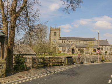 A street view with wooden benches and a road near a stone church with a clock tower at Doe Foot Cottage in Ingleton
