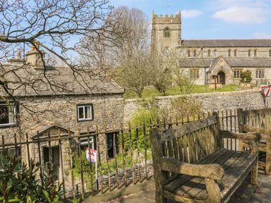 Stone house with garden and iron fence next to wooden benches and church in background at Doe Foot Cottage in Ingleton