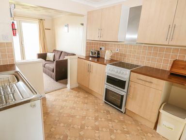 A kitchen with cupboards a stove and toaster opening to a living area with a brown sofa at Surf in Hunstanton