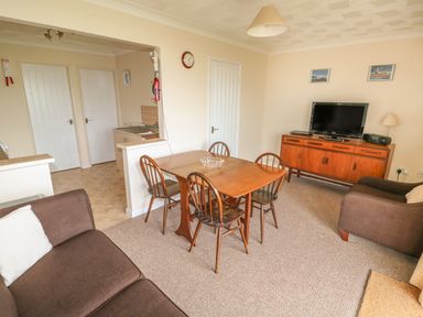 A living room with a wooden dining table and four chairs a television on a wooden cabinet a brown sofa and armchair and an open doorway to a kitchen at Surf in Hunstanton