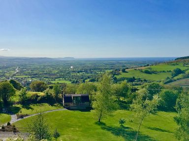 A landscape view of green fields trees and a small cabin with hills and the sea in the background at Idris Pod in St Asaph