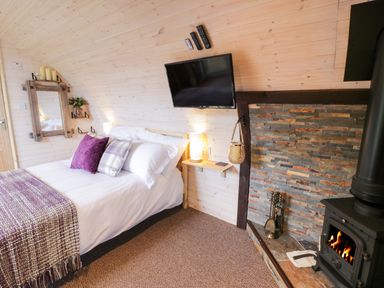 A bedroom with a bed pillows and blanket a wall-mounted television and a fireplace with fire at Gelert Pod in St Asaph