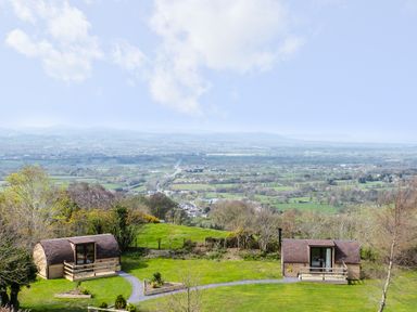 Two small wooden pods with decks on a grassy hill overlooking a valley with fields and trees at Gelert Pod in St Asaph