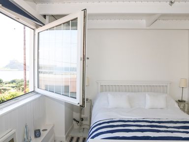 A bedroom with an open window showing a view outside and a bed with pillows and a striped blanket at Ner Sea in Marazion