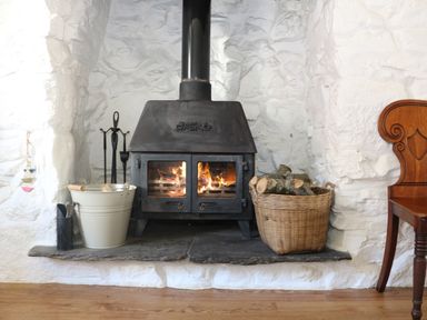 A wood burning stove with fire inside a white stone fireplace with a basket of logs a white bucket with tools and a wooden chair next to it at Ner Sea in Marazion