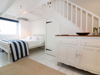 A bedroom with a white bed and striped blanket next to a white cabinet under a staircase at Ner Sea in Marazion