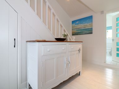 A white wooden cabinet under a staircase with a bowl and flowers on top and a painting on the wall at Ner Sea in Marazion