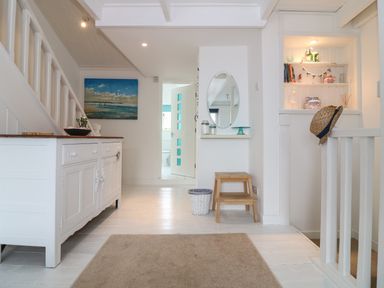 A hallway with a white cabinet under a staircase a painting on the wall a round mirror a small wooden stool and an open doorway at Ner Sea in Marazion