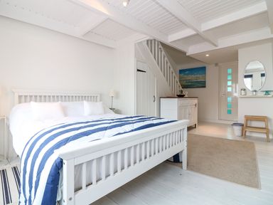 A bedroom with a white wooden bed striped blue and white bedding a white dresser under stairs a round mirror on wall and a small bench at Ner Sea in Marazion