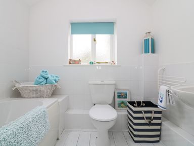 A bathroom with a toilet, bathtub, wicker basket with towels, striped laundry basket, and small window with blue blind at Ner Sea in Marazion
