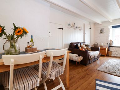 A living room with a wooden table and chairs sunflowers on the table a brown leather sofa and a window with a ledge at Ner Sea in Marazion