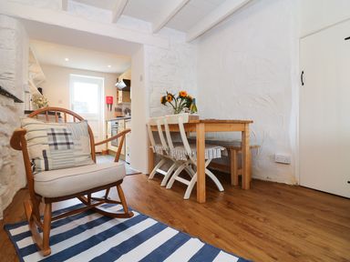 A dining area with wooden table and chairs next to a kitchen and a rocking chair on a striped rug at Ner Sea in Marazion