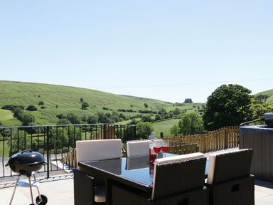 An outdoor patio with a glass table set and a barbecue grill overlooking green hills at 1 Beacon View Barn in Beguildy near Knighton