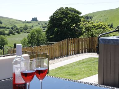 A garden with a wooden fence overlooking green hills with two glasses of red wine and a bottle on a table near a hot tub at 1 Beacon View Barn Beguildy near Knighton