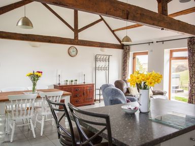 A dining area with white chairs around a wooden table and flowers in a vase a living area with armchairs and large windows at 1 Beacon View Barn in Beguildy near Knighton