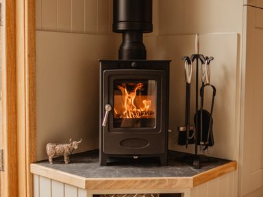 A wood burning stove with fire inside on a corner countertop with fireplace tools and a decorative animal figure at Moelfre - Shepherds Hut in Llanbedr