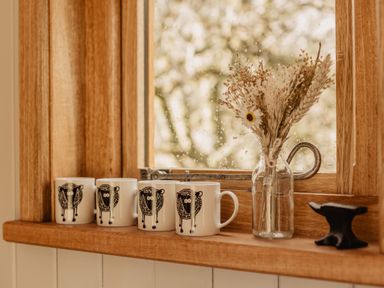 A wooden window sill with four mugs decorated with sheep illustrations a glass vase with dried flowers and a small black anvil at Moelfre - Shepherds Hut in Llanbedr
