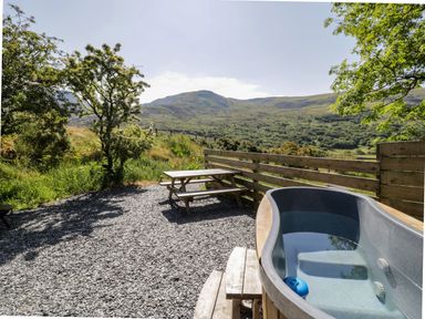 An outdoor area with a hot tub and a wooden picnic table with benches surrounded by a wooden fence and trees at Moelfre - Shepherds Hut in Llanbedr