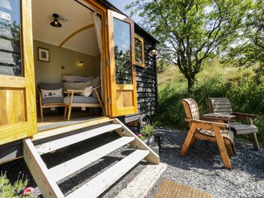 A small cabin with open doors showing two chairs inside and two wooden chairs outside near trees at Moelfre - Shepherds Hut in Llanbedr