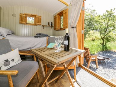 A wooden table with wine bottle and glasses next to a gray armchair and bed in a small room with wooden walls and open door to garden at Moelfre - Shepherds Hut Llanbedr