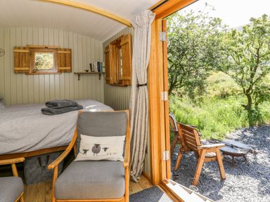 A small bedroom with a bed towels an armchair with a pillow wooden framed windows and a door opening to an outdoor seating area with chairs and trees at Moelfre - Shepherds Hut in Llanbedr