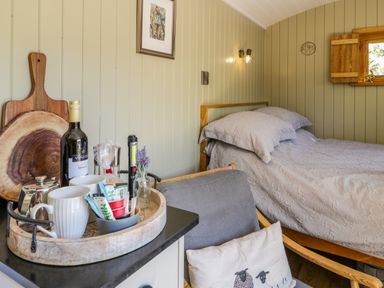 A bedroom with a wooden bed and gray bedding next to a chair with a cushion and a tray with mugs and a bottle of wine at Moelfre - Shepherds Hut in Llanbedr