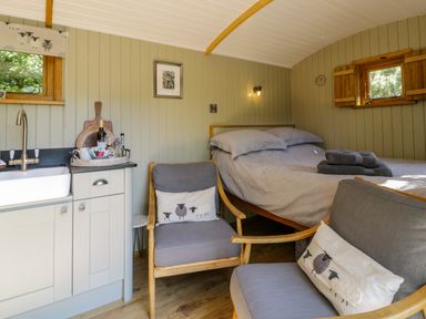 A small room with a bed next to two chairs with sheep cushions and a kitchenette with a sink at Moelfre - Shepherds Hut in Llanbedr
