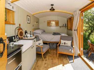 A small room with a bed at the back two chairs with cushions a sink a countertop and wooden door open to outside at Moelfre - Shepherds Hut in Llanbedr