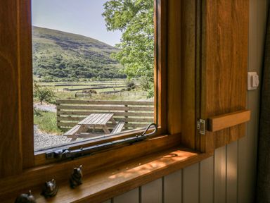 A wooden window with a latch looking out to a wooden picnic table and a green hill at Moelfre - Shepherds Hut in Llanbedr