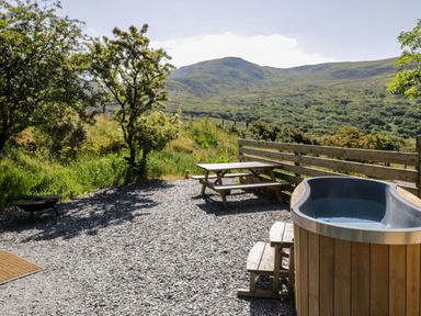 An outdoor seating area with a wooden hot tub a picnic table and a fire pit with mountains in the background at Moelfre - Shepherds Hut in Llanbedr