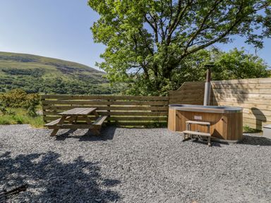 An outdoor patio area with a wooden picnic table and a wooden hot tub with steps next to a wooden fence and trees at Moelfre - Shepherds Hut in Llanbedr