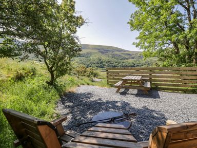 A wooden picnic table and two wooden chairs with a fire pit on a gravel area surrounded by trees and hills at Moelfre - Shepherds Hut in Llanbedr